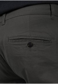 I pantaloni grigi scuri presentano una tasca posteriore con chiusura a bottone e un'accademia in tessuto a righe. Il tessuto appare liscio con una texture minima.