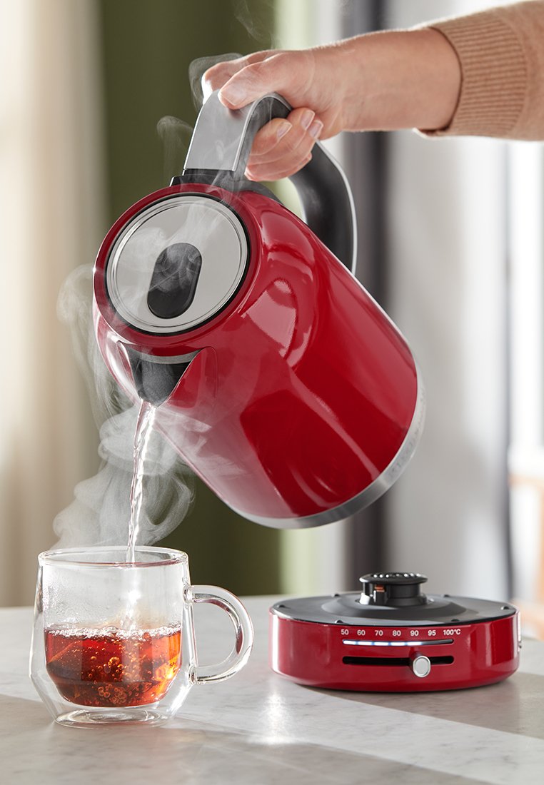 Röd elektrisk vattenkokare med en spout i rostfritt stål, hällande hett vatten i en klar glas kopp fylld med bryggt te. Elektrisk bas synlig.