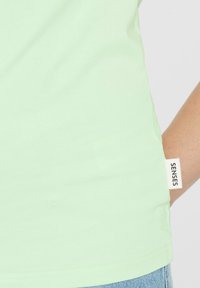 Lichtgrünes Baumwoll-T-Shirt mit einer glatten Textur, kurzen Ärmeln und einem sichtbaren "SENSES"-Etikett an der Seitennaht.