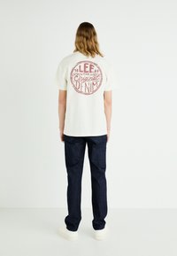 White cotton t-shirt with red graphic print featuring "LEE Original Denim" on the back, paired with dark denim jeans and white sneakers.