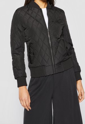 Femme portant une veste bomber matelassée noire avec fermeture éclair à l'avant et poignets côtelés, par-dessus un top blanc, associée à un pantalon noir ample.