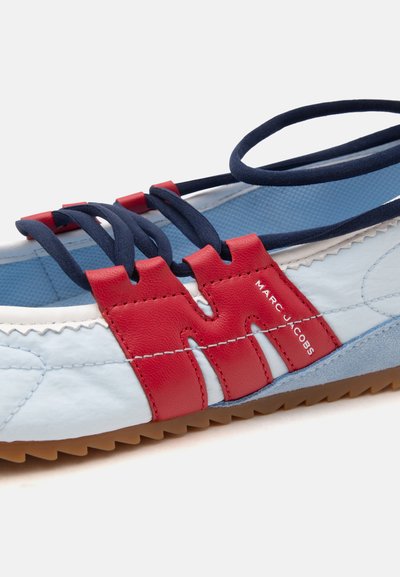 Gros plan d'une basket bleue et blanche avec un détail en cuir rouge et des lacets élastiques bleu marine, avec le nom de la marque "Marc Jacobs" sur le côté.
