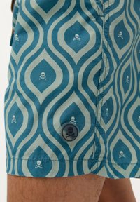 Shorts de bain turquoise avec un motif de vagues et des designs de crânes et os croisés blancs. Comprend un bouton avec un graphique de crâne.