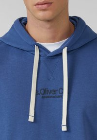 Uomo che indossa una felpa blu con cappuccio, con stringhe bianche e la scritta nera "s.Oliver C." parzialmente visibile sul petto sopra una camicia bianca.