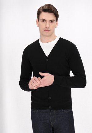 Cardigan nero realizzato in tessuto a maglia, con scollo a V, maniche lunghe e sei bottoni frontali. Abbinato a una camicia bianca e jeans.