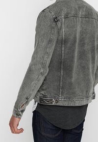 Šedá džínová bunda s opotřebovaným vzhledem, zapínáním na knoflíky na manžetách a dvěma náprsními kapsami. Má standardní límec a mírně přiléhavý střih.