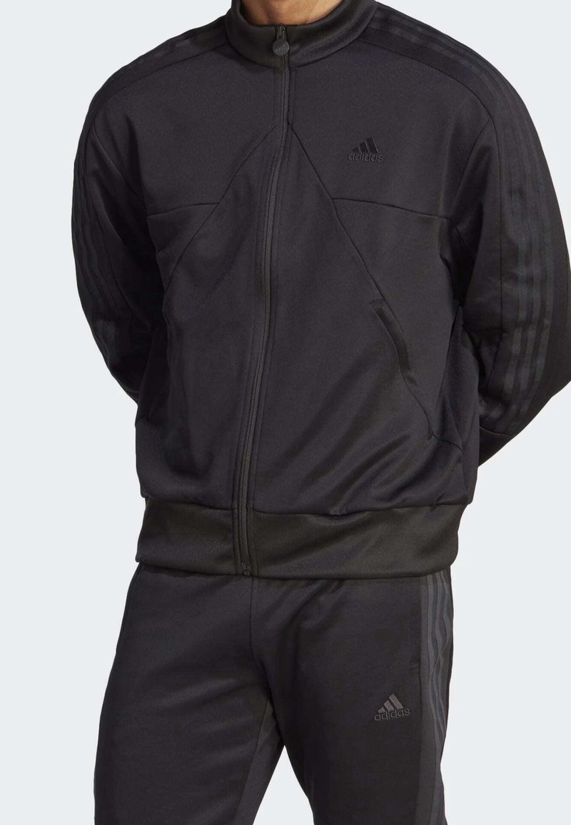 Svart träningsdräkt med en zip-up jacka som har strukturerade paneler och tre-rands accenter på ärmarna och byxorna.