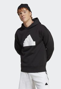 Black hoodie with a large white three-stripe logo on the front. Features a drawstring hood and ribbed cuffs. Worn with white shorts.