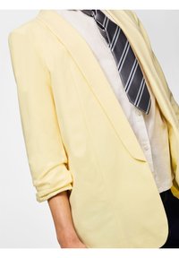 Personne portant un blazer jaune pâle aux manches retroussées, une chemise blanche boutonnée et une cravate rayée gris foncé sur un fond blanc.