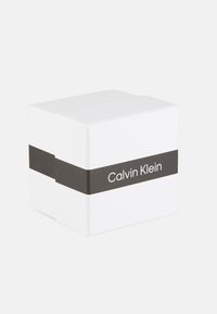 Boîte carrée blanche avec une finition mate, présentant un bandeau en papier noir sur lequel est affiché le logo "Calvin Klein" en texte blanc.