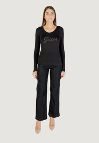 Maglietta nera a maniche lunghe con logo "Guess" testurizzato in argento. Abbinata a jeans flared di denim scuro e scarpe col tacco color nude.