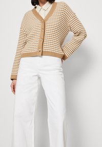 Beige-weiß gestreifte, kurze Strickjacke mit V-Ausschnitt und fünf Knöpfen, kombiniert mit weit geschnittenen weißen Hosen. Die Strickstruktur ist sichtbar.