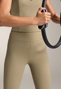 Beige szövetű sportfelső és hozzá illő magas derekú leggings, melyekben a kezek fekete fitness gyűrűt szorítanak, bemutatva az anyag bordázott mintázatát.