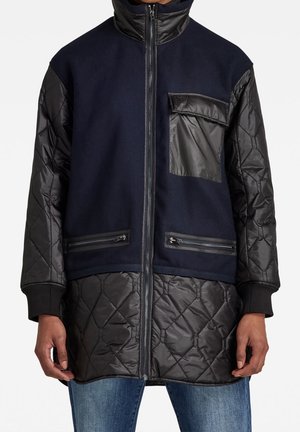 Person trägt eine lange, gesteppte schwarze und marineblaue Jacke mit Reißverschlusstaschen, vorne mit Reißverschluss und einer Klappentasche auf der Brust, kombiniert mit blauen Jeans.