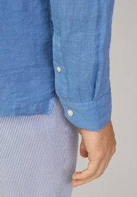 Camicia a maniche lunghe in lino blu con polsini abbottonati, caratterizzata da una texture liscia. Abbinata a pantaloni a righe, mostrando una mano che afferra la camicia.