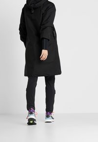 Abrigo negro impermeable con capucha, mangas holgadas y bolsillos con cremallera, combinado con pantalones ajustados negros y zapatillas deportivas multicolor.