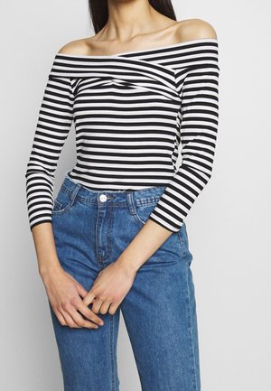 Vrouw draagt een zwart-wit gestreept off-shoulder top met lange mouwen en een hoge blauwe spijkerbroek, handen gevouwen voor zich.