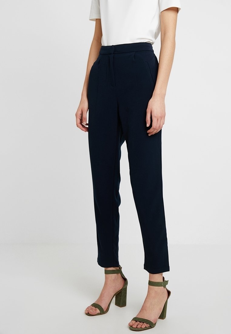 Femme portant un pantalon bleu marine ajusté longueur cheville, un haut blanc à manches courtes et des sandales à talons hauts vert olive avec des lanières aux chevilles.