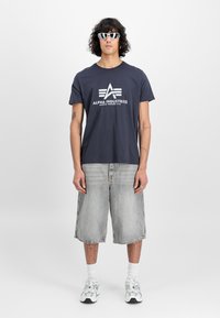 T-paita, tummansininen puuvillasta, jossa on valkoinen "ALPHA INDUSTRIES" -logo, yhdistettynä vaaleanharmaisiin farkkushortseihin, valkoisiin sukkiin ja harmaisiin lenkkareihin.