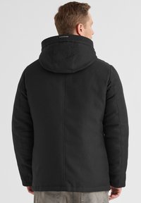 Schwarze isolierte Jacke mit Kapuze, die eine glatte Oberfläche und ein strukturiertes Design aufweist. Die Rückansicht zeigt Nahtdetails und das Logo am Kragen.