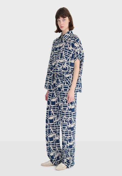 Femme portant une chemise ample assortie et un pantalon large bleu marine et blanc à motifs, avec des chaussures blanches sans lacets, debout devant un fond clair.