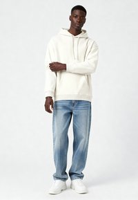 Sweat à capuche beige clair avec une poche frontale, une texture douce et une capuche à cordon, associé à un jean bleu délavé et des baskets blanches.