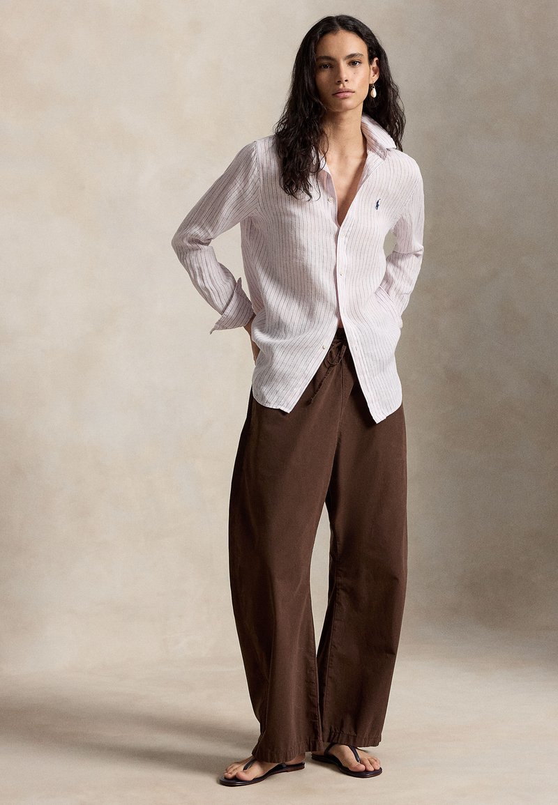 Femme aux longs cheveux bruns portant une chemise à rayures claire boutonnée, un pantalon large marron et des tongs noires, debout les mains dans les poches.