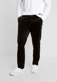 Man wearing dark brown corduroy pants, white long-sleeve shirt, white sneakers, standing with hands in pockets on plain background.