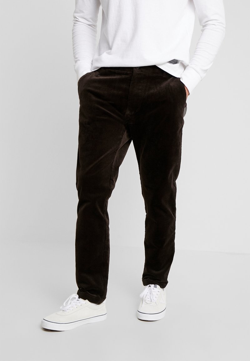 Man wearing dark brown corduroy pants, white long-sleeve shirt, white sneakers, standing with hands in pockets on plain background.