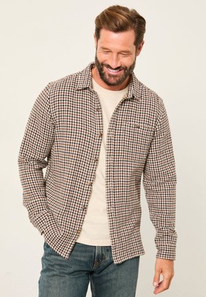 Smiling man with beard wearing a brown and white houndstooth button-up shirt over a beige t-shirt and blue jeans, standing against a plain background.