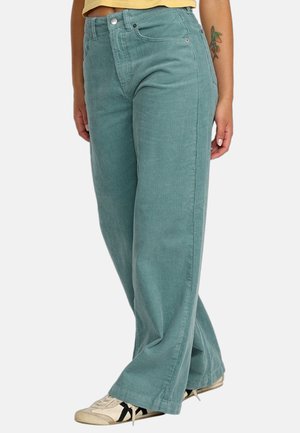Personne portant un pantalon large en velours côtelé turquoise, des baskets beiges avec des rayures noires, et un haut jaune, debout jambes croisées.