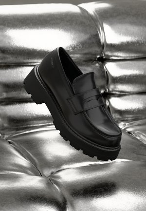 Mocasines negros brillantes con suela gruesa negra, con una correa frontal y punta redondeada, exhibidos sobre un fondo metálico plateado.