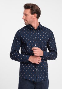 Marineblauwe shirt met kleine, lichtoranje patronen. Lange mouwen, knoopsluiting en een gladde textuur, afgewerkt met knopen aan de voorkant.