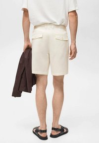 Cremefarvede shorts med to baglommer, bærer en lys skjorte og en brun jakke, parret med sorte sandaler.