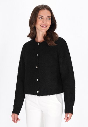 Cardigan nero lavorato a maglia con scollo rotondo, maniche lunghe e bottoni argento. La texture è morbida e pelosetta, con una vestibilità corta.