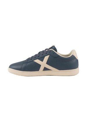 Zapatilla deportiva azul oscuro con suela beige, cordones y gran logo X en el lateral, mostrada desde el lado exterior sobre un fondo blanco.