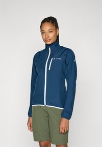 Marineblå lett jakke med høy krage, hvit glidelås og elastiske ermer. Kombinert med olivengrønne shorts for et utendørsutseende.