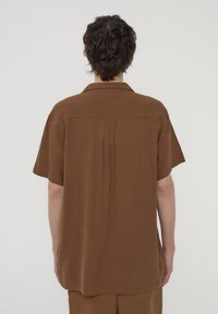 Camicia marrone a maniche corte realizzata in un tessuto strutturato con una vestibilità rilassata, dotata di un dietro con yoke e dettaglio plissettato, colletto e orlo arrotondato.