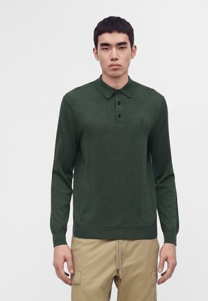 Jeune homme portant un polo à manches longues vert foncé et un pantalon cargo beige, debout devant un fond clair uni.