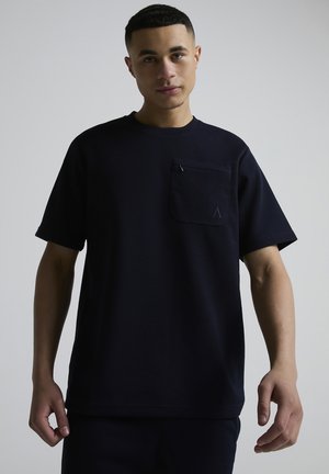 Ung mann med kort hår iført en svart strukturert kortermet skjorte med en glidelåslomme på brystet, stående foran en ensfarget grå bakgrunn.