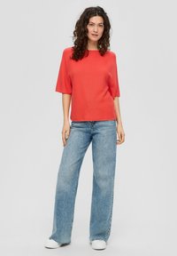 Rotes kurzärmliges Oberteil aus strukturierter Stoff, kombiniert mit hellblauen weit geschnittenen Jeans und weißen Sneakern, was einen lässigen Look ergibt.