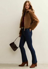 Brązowa kurtka z krótkim futerkiem ze sztucznego futra, bordowa koszula, ciemnoniebieskie rozkloszowane dżinsy i brązowe zamszowe botki. Trzyma ciemną plecioną torebkę.