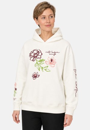 Vrouw draagt een witte hoodie met bloemmotieven en tekst "roots before wings" en "The earth" op de mouwen, staat met ontspannen handen.