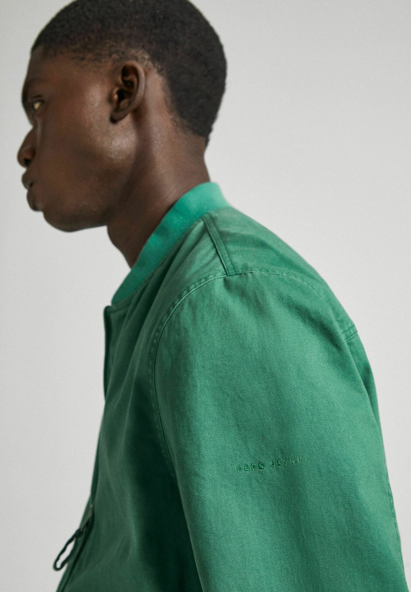 Chaqueta bomber verde con un acabado suave, cuello acanalado y logotipo en relieve en la manga. Cuenta con cierre de cremallera y bolsillos laterales.