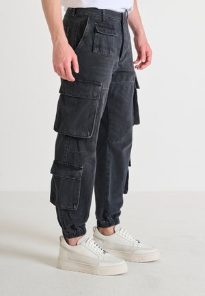 Pantaloni cargo neri in denim, dotati di numerose tasche grandi, fondo svasato e vita elasticizzata, abbinati a sneakers bianche.