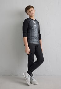 Băiat îmbrăcat într-un costum sport negru și adidași albi, sprijinit de un perete gri cu textură, stând cu un picior încrucișat peste celălalt.