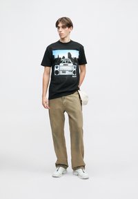 Juodas medvilninis marškinėlis su grafika, vaizduojančia boomboxą ir miesto panoramą. Derinamas su smėlio spalvos kelnėmis ir baltais sportbačiais. Modelis stovi prie neutralaus fono.