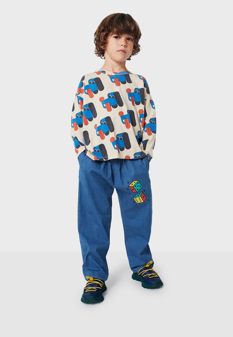 Long-sleeve cream shirt with multicolor vehicle print, paired with blue denim pants featuring a dice graphic, worn with multicolored shoes.