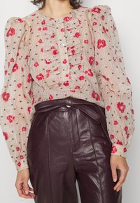 Helle beige Bluse mit roten Blumen- und braunen Punkte-Mustern, ausgestattet mit Rüschendetails und drei dekorativen Knöpfen. Lange Puffärmel.