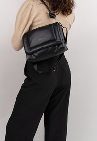 Donna che indossa un maglione beige e pantaloni neri, mentre porta una borsa a spalla in pelle nera con la scritta dorata "EXPATRIÉ" su uno sfondo chiaro.